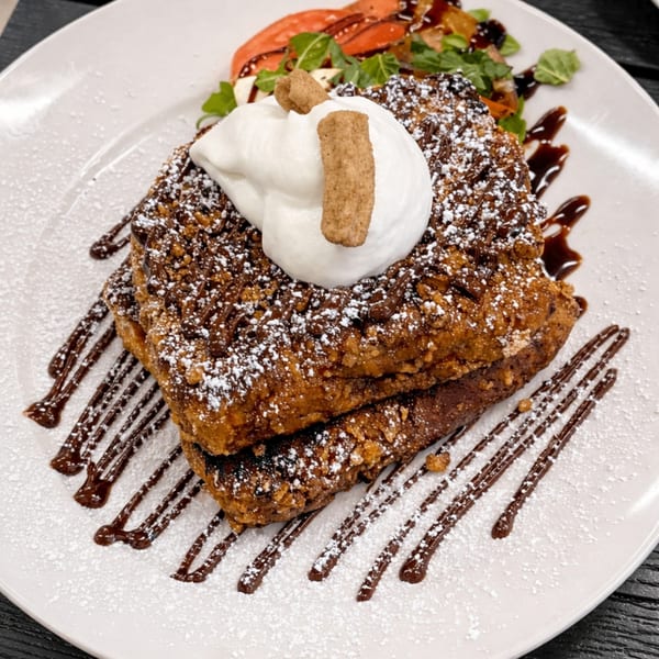 Two pieces of French toast topped with whipped cream, chocolate drizzle, and powdered sugar.