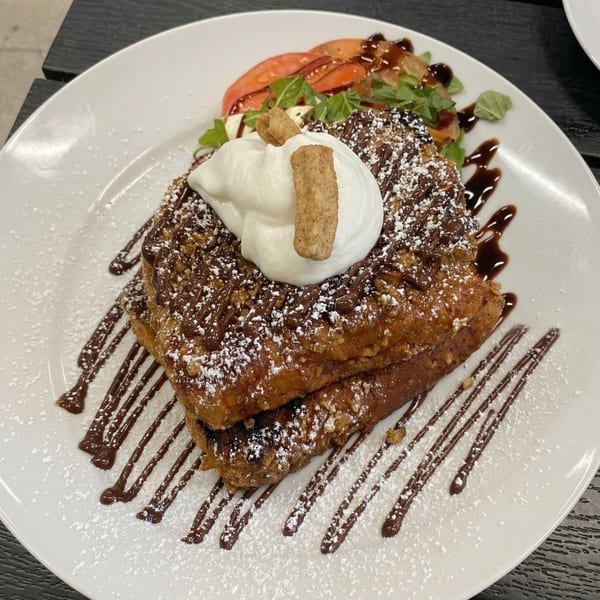 Two pieces of French toast topped with whipped cream, chocolate drizzle, and powdered sugar.